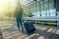 Ragazza di spalle che cammina verso sinistra su un marciapiede fuori da un aeroporto, ha uno zaino in spalla e un trolley, indossa pantaloni lunghi e t-shirt.